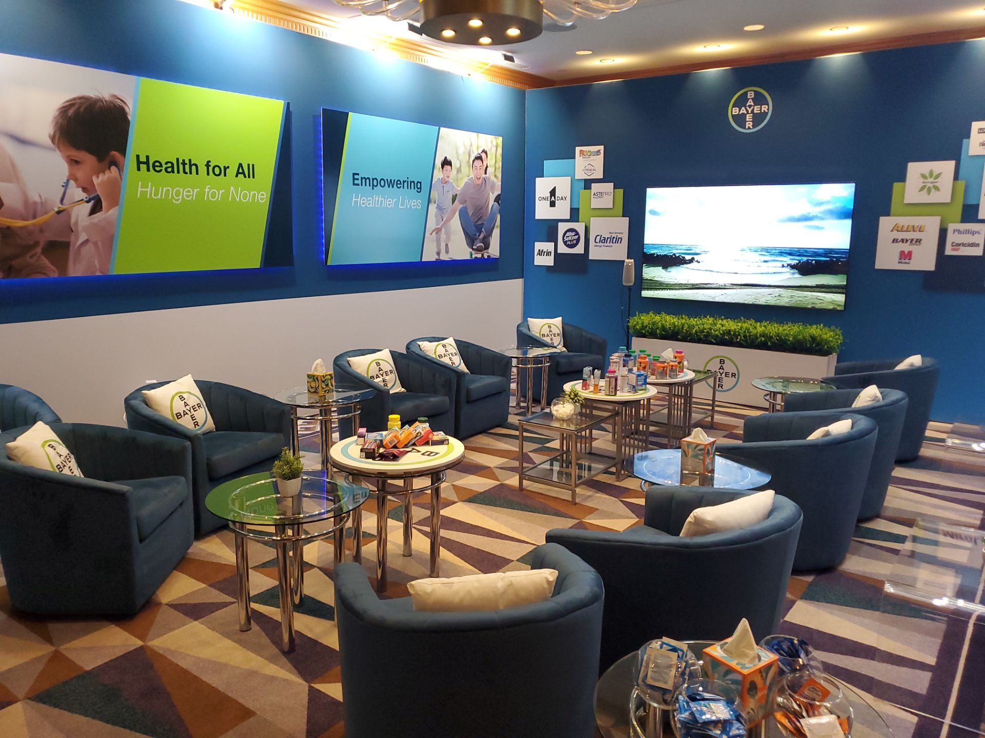 Annual Meeting suite — wide shot showing branded lounge with Bayer pillows, product shelves, and tiered display